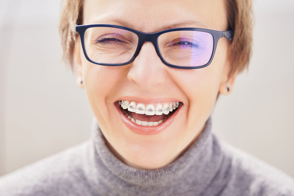 Stylish and beautiful young blonde woman in glasses and a gray sweater in braces and smiling on a white background, dentist orthodontist concept