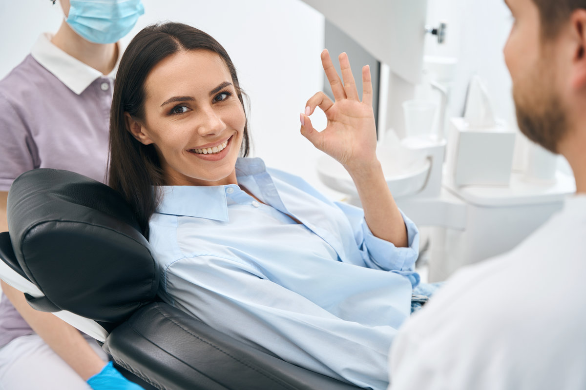 Positive woman client of stomatological clinic showing ok sign gesture sitting on orthodontic chair, enjoying good service in private dental clinic, routine teeth check-up